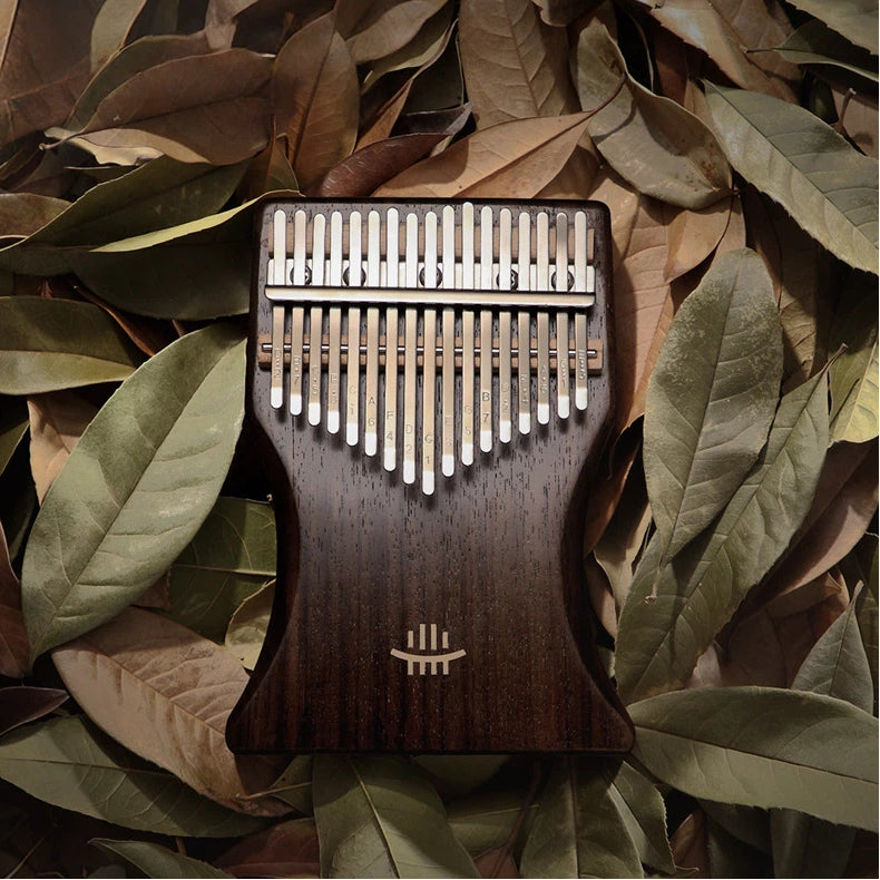 A 34-key double-layer black walnut kalimba by Hluru, shot from a top-down view on a bed of dried leaves, showcasing its exquisite wood grain and engraved tines.