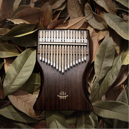 A 34-key double-layer black walnut kalimba by Hluru, shot from a top-down view on a bed of dried leaves, showcasing its exquisite wood grain and engraved tines.