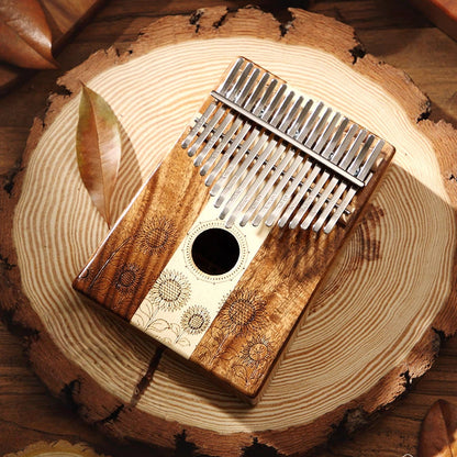 A 17-key hollow-body kalimba by MoralGain, featuring intricate sunflower engravings, a unique light-and-dark wood grain patchwork design, and a central sound hole, displayed on a natural wood ring base.