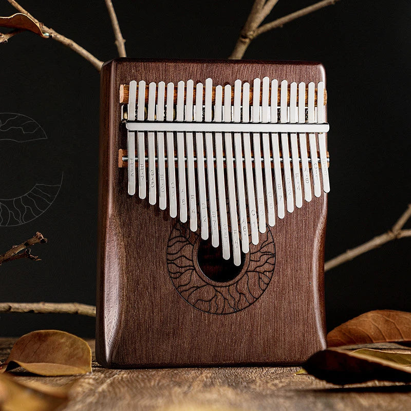 A 21-key kalimba thumb piano made of dark wood with an engraved eye-shaped pattern, featuring stainless steel tines with etched musical notes, set against a background of dried leaves.