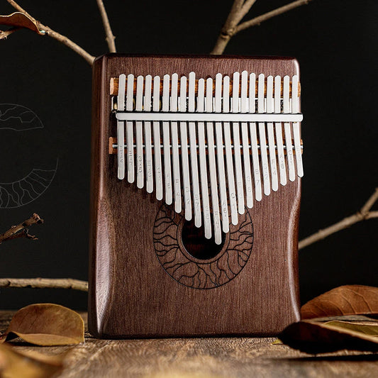 A 21-key kalimba thumb piano made of dark wood with an engraved eye-shaped pattern, featuring stainless steel tines with etched musical notes, set against a background of dried leaves.
